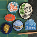 A collection of iron on embroidered patches. From Left to Right: A be kind patch with floral script, an oval patch of an eagle claw and olive brand that says "hold fast", a patch that says chasing wonder, a navy blue patch featuring a golden anatomical heart, a patch with a dog and human holding hands and text saying "friend to strays", and a die cut cloud patch with dreamer script along the bottom