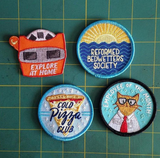 From left to right: An orange viewfinder patch that says "explore at home", a circle sunrise patch that says "reformed bedwetters society", a blue circle patch with a slice of pizza in the center and text over it that says, "cold pizza club", and a teal circle patch with an orange cat named Bill with text above stating, "employee of the month"