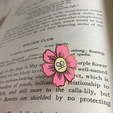 An enamel pin with a butterfly back in the shape of a pink flower with a very silly indifferent face in the center on top of the text of a book