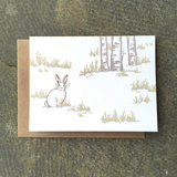 A greeting card showing a snow covered ground with patches of grass and a white rabbit.