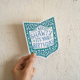Die-cut shield-shape greeting card and envelope. Card reads "go shawty it's your birthday" in ornate, hand written, blue type. Surrounded by an ornate border in shades of blue being held in front of a white wood wall