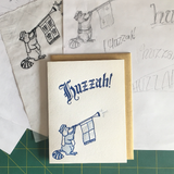 Greeting card and kraft paper envelope. Text in old-timey blackletter reads, "huzzah!" Illustration below of raccoon in medieval garb and heralding trumpet that says "toot!"