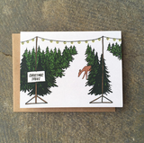 A greeting card featuring rows of Christmas trees for sale with twinkle lights and a deer meandering in between.